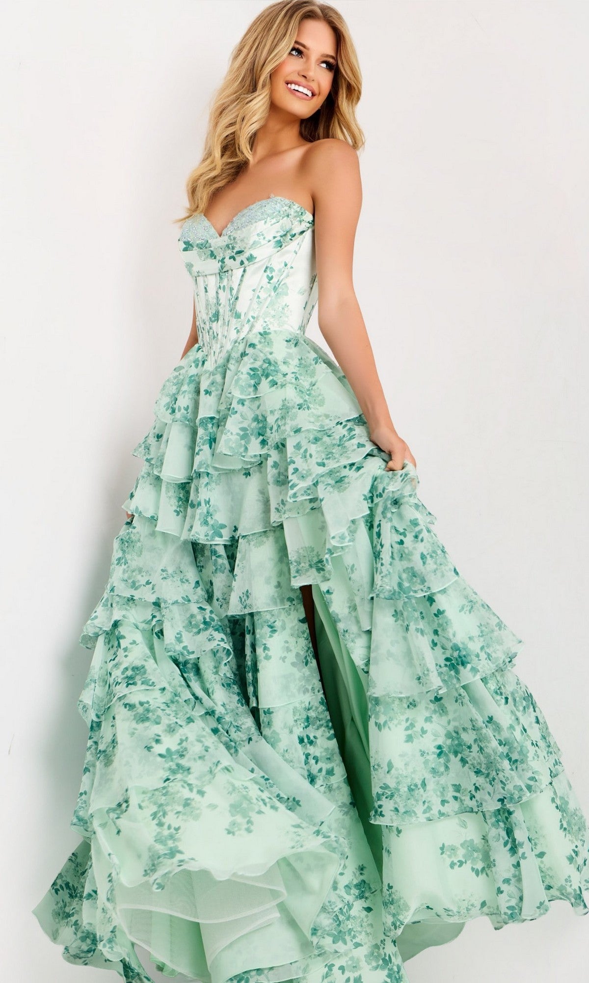 A woman smiles in the Jovani 48772 long prom dress, featuring a strapless corset bodice, tiered mint green ruffle layers, and floral print, standing against a plain light background.